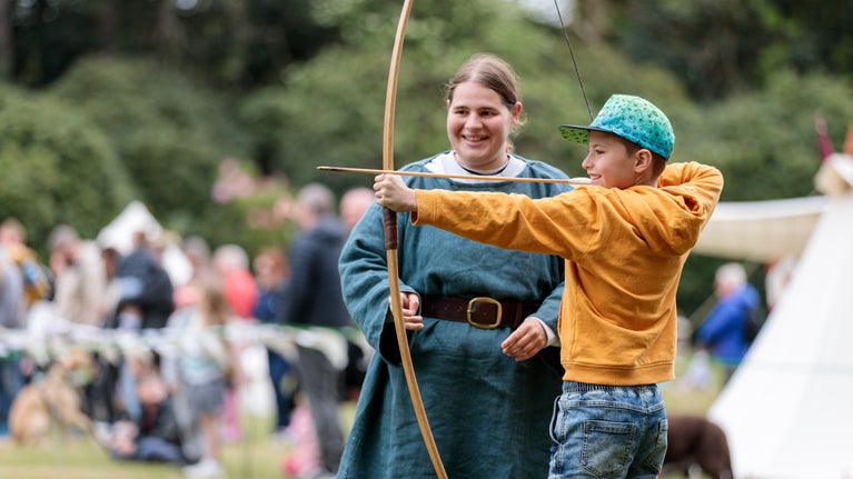 Two people at an outdoor event practice archery; one in a yellow hoodie and blue cap aims a bow, while another in green stands nearby. Blurred crowd and tents are in the background.